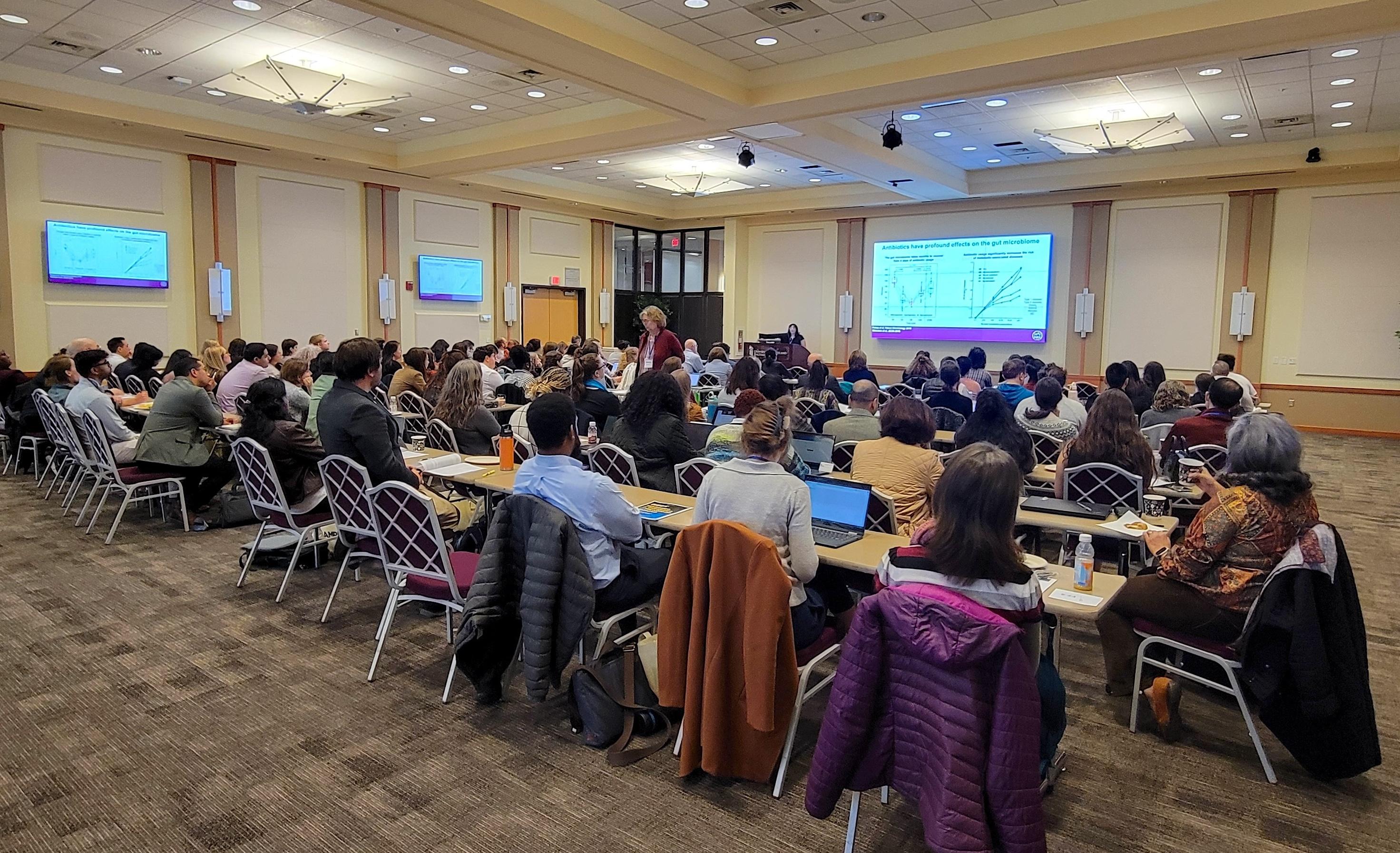 A large audience attends a microbiome research seminar, engaging with a speaker presenting on antibiotics' effects on the gut microbiome.
